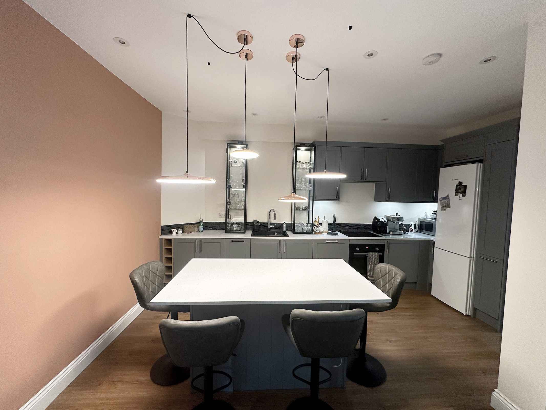 Wide view of an open-plan grey kitchen with a large white island, bar seating and suspended pendant lights